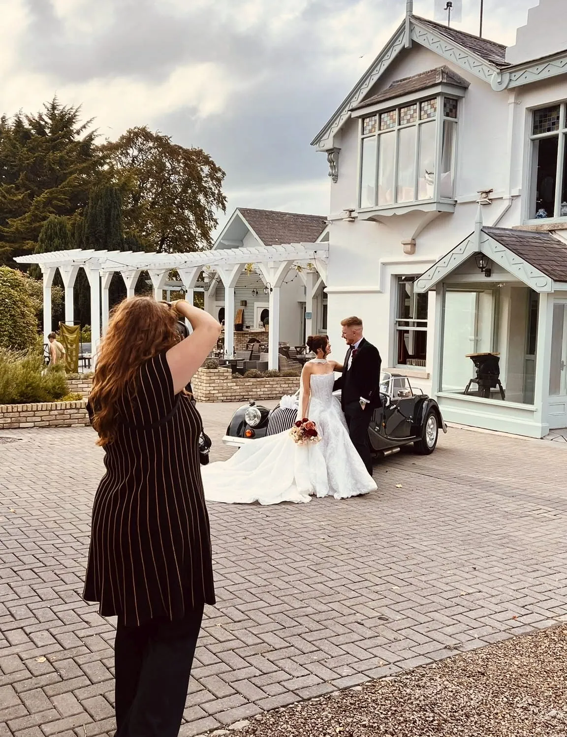 Lucy photographing a couple beside a vintage car outside a venue