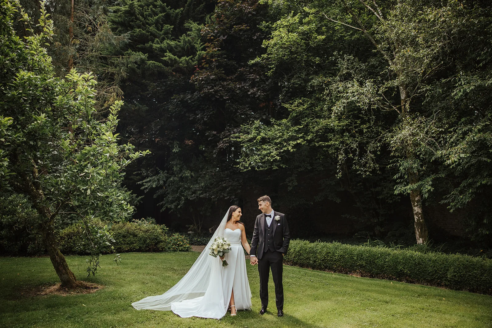 A couple hand in hand on a lawn in front of tall trees